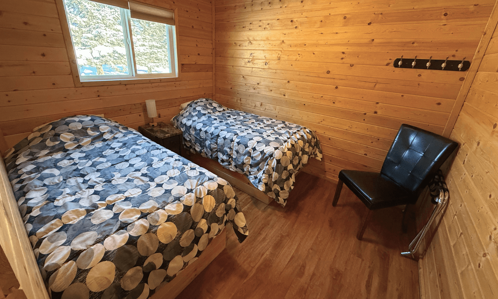 Cozy cabin bedroom with two single beds, blue-and-white duvets, a black leather chair between, window above, and a wall coat rack.