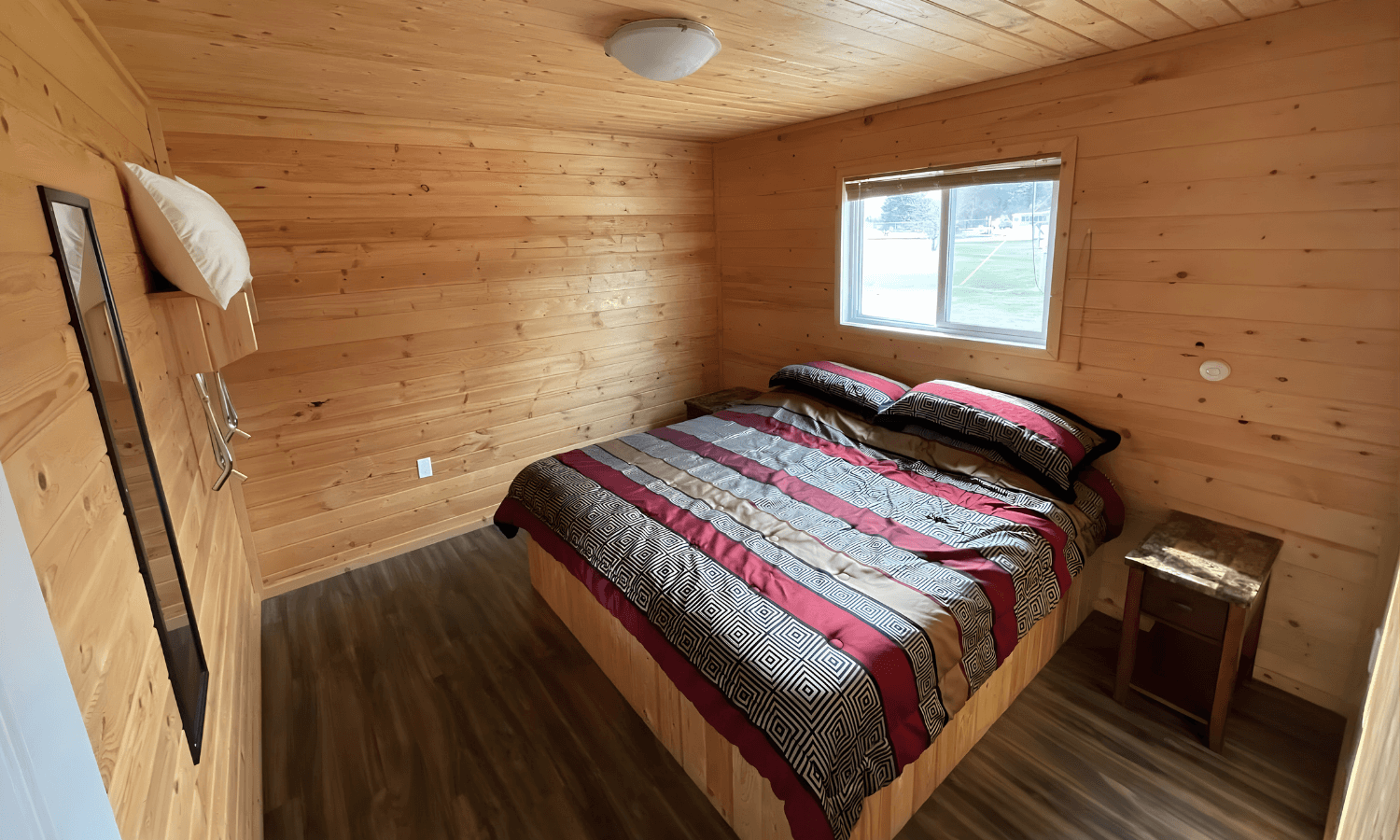 wood-paneled bedroom with a bed, striped blanket, window, and wooden floor.