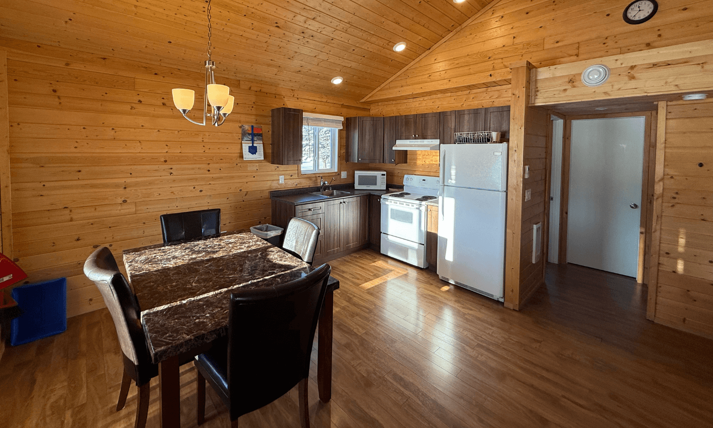 cabin interior: dining table with chairs, small kitchen with brown cabinets and a white fridge, wood-paneled walls and ceiling, wooden floor.