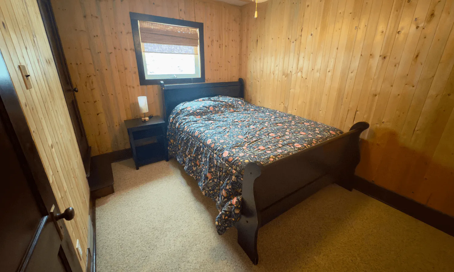 Rustic cabin bedroom with wooden paneled walls, a twin bed with blue patterned bedding, and a small window.