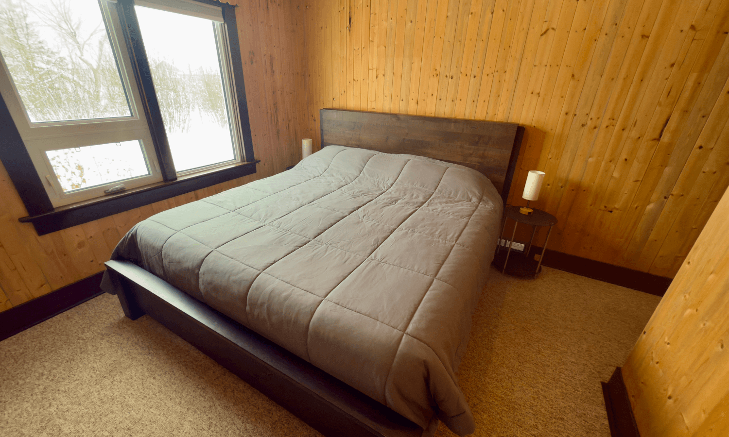 bedroom with warm wooden walls, a gray bed with simple bedding, and windows.