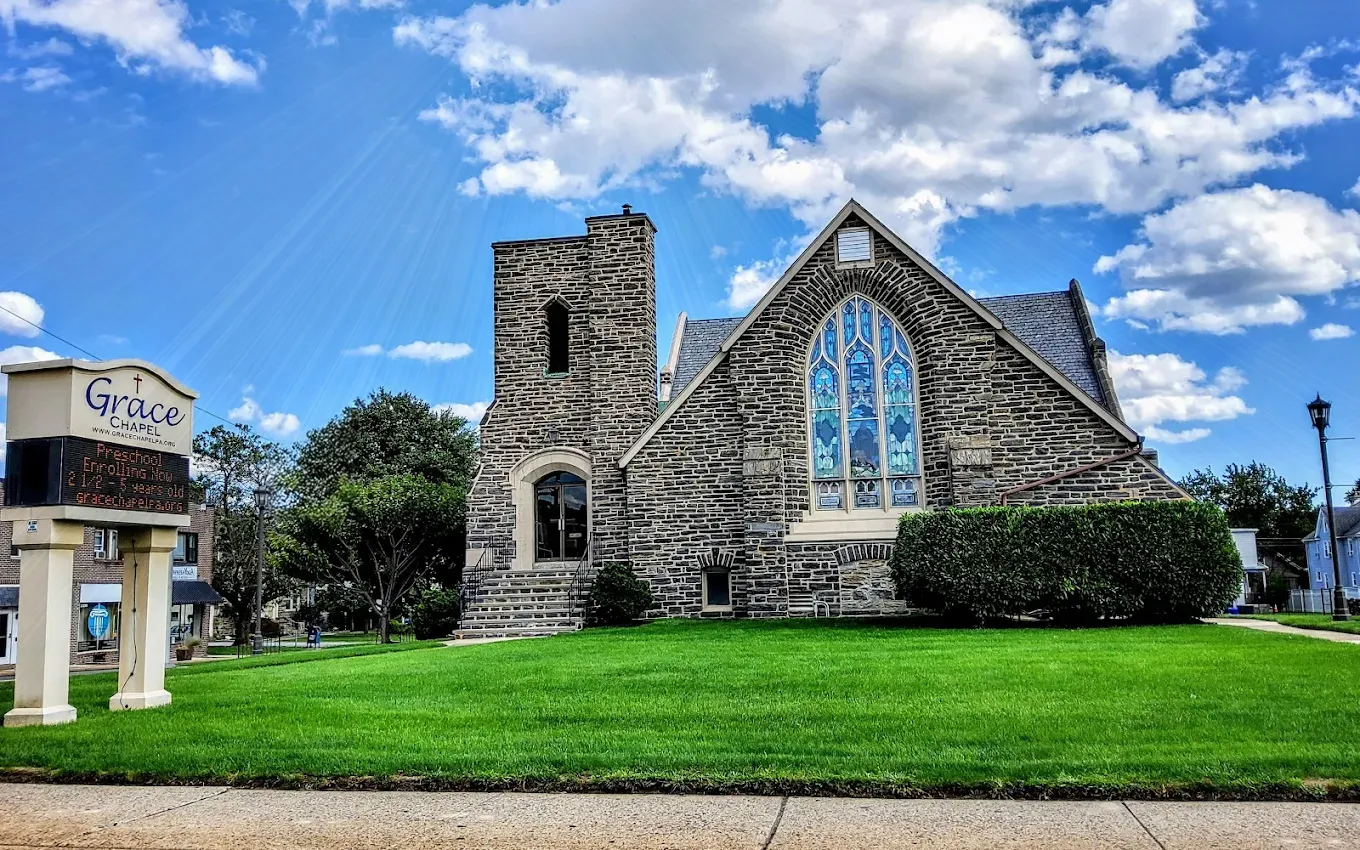 Picture of Grace Chapel located in Havertown, PA. Church is gray color stone with stain glass windows and a door to the left. The church is sitting on a plush green lawn with the Grace Chapel sign infront to the left of the church