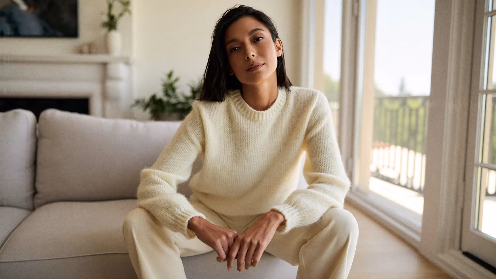 A woman seated beside a bright window in a cream knit sweater, with healthy, well-cared-for skin