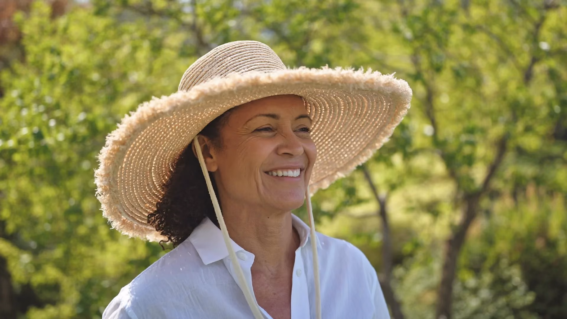A woman in a wide-brimmed sun hat smiling outdoors in a bright garden