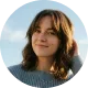 Smiling woman with shoulder-length brown hair and bangs, wearing a blue checkered shirt, outdoors with a cloudy sky background.
