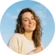 Smiling woman with curly hair wearing a beige top against a blue sky background.