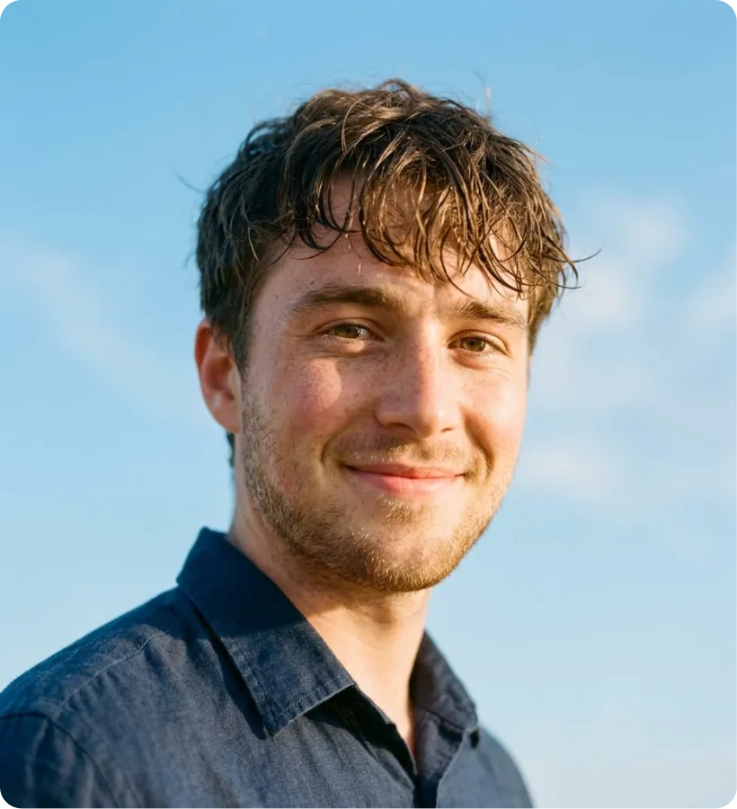 Smiling young man with tousled brown hair and light facial hair wearing a navy blue shirt against a clear blue sky background.