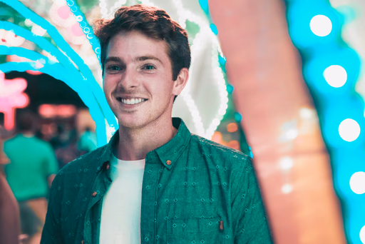 Smiling young man in a green shirt standing in a vibrant, colorful setting with blurred lights in the background.