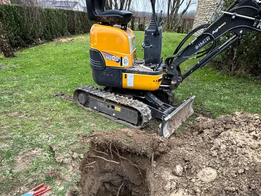 Yellow mini excavator digging a hole in a grassy yard near a hedge and stone wall.