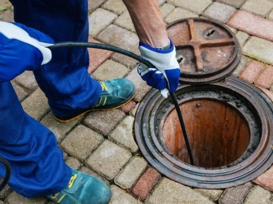 Worker in blue protective clothing using a tool to clear a drain or sewer covered by a metal manhole cover on a paved surface.