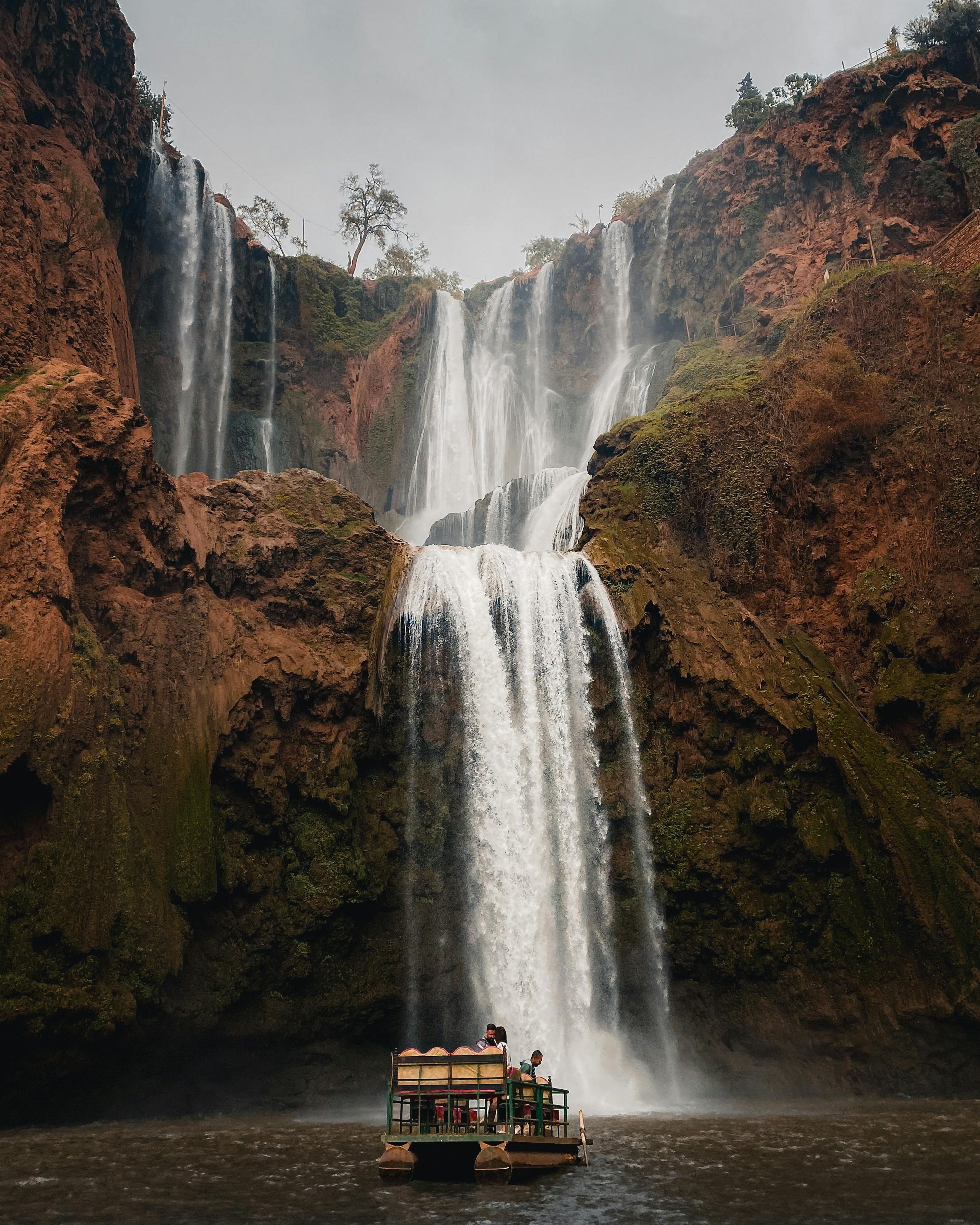 People sitting on a small raft approaching a large multi-tiered waterfall surrounded by rocky cliffs.