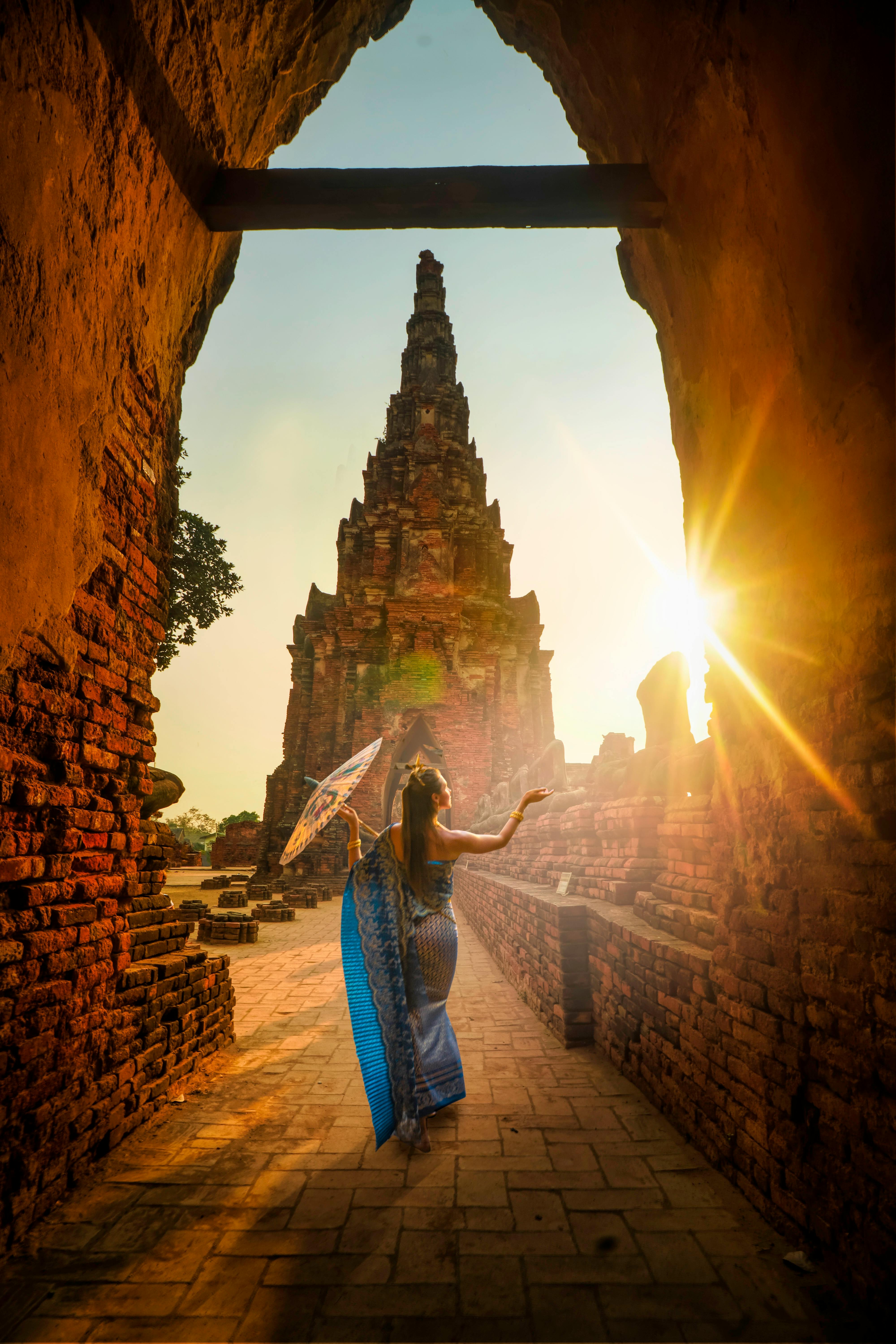 Woman in traditional blue dress holding a parasol walking through ancient brick ruins toward a tall temple at sunset.