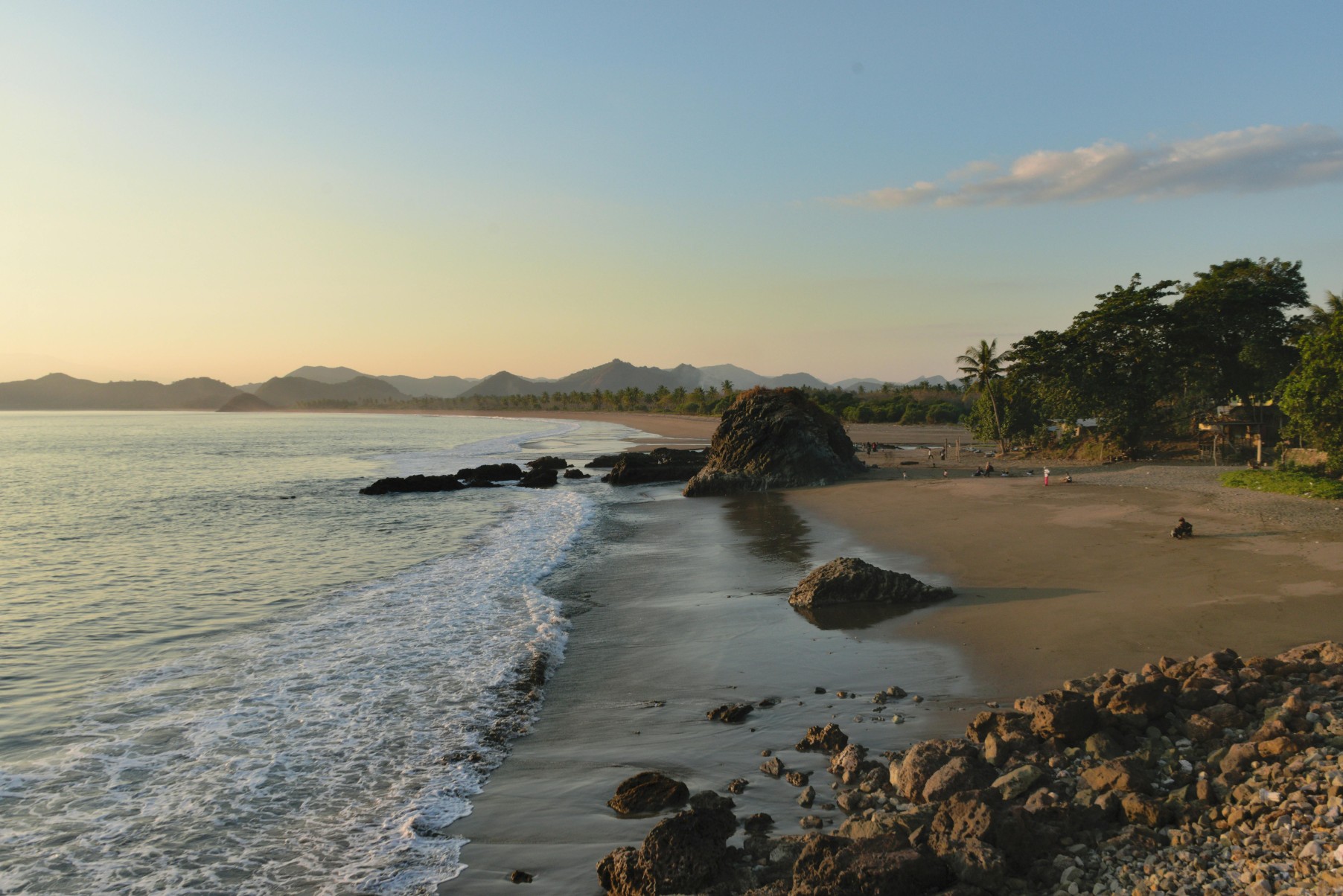 A peaceful sunset view over a rugged Lombok beach with gentle waves and rock formations.