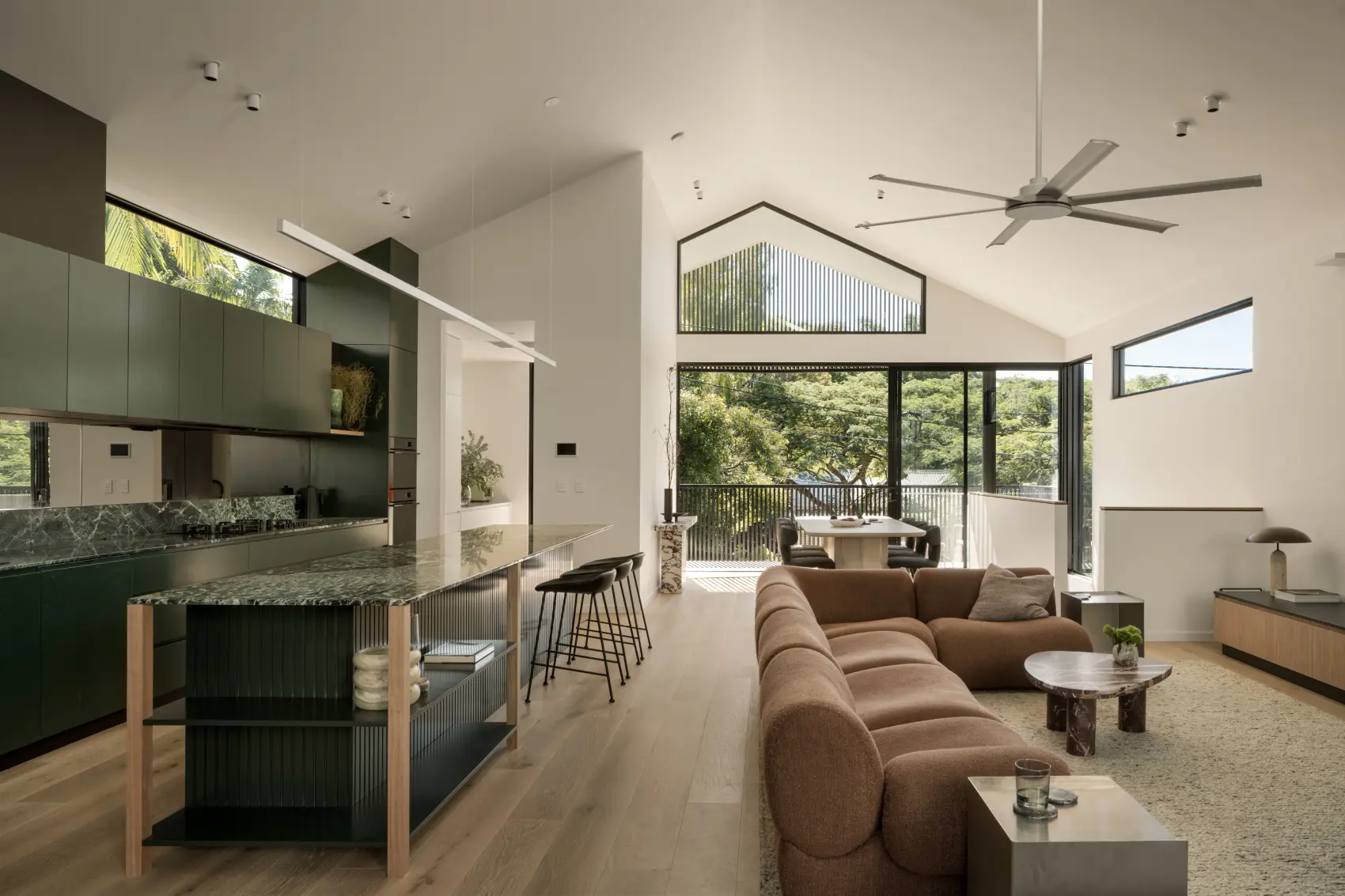 Modern open living room and kitchen with large windows, brown sofa, island, and dining table with chairs.