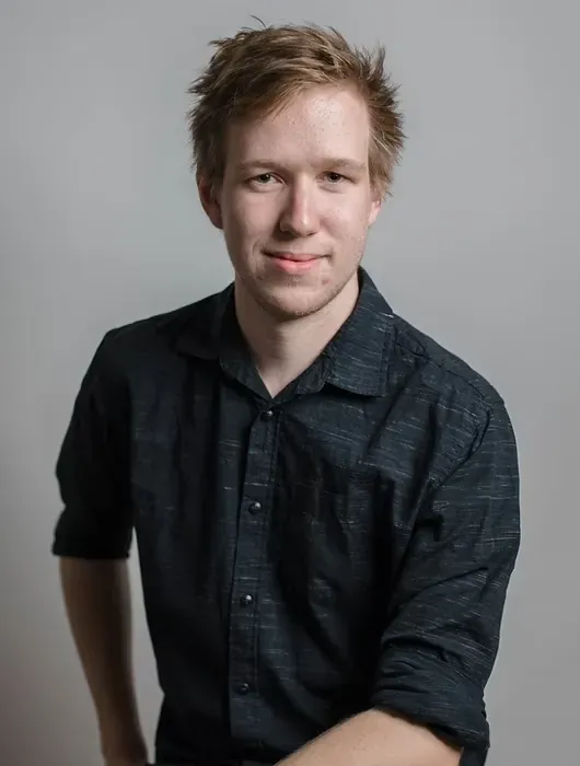 Young man with tousled blond hair wearing a dark button-up shirt against a plain gray background.