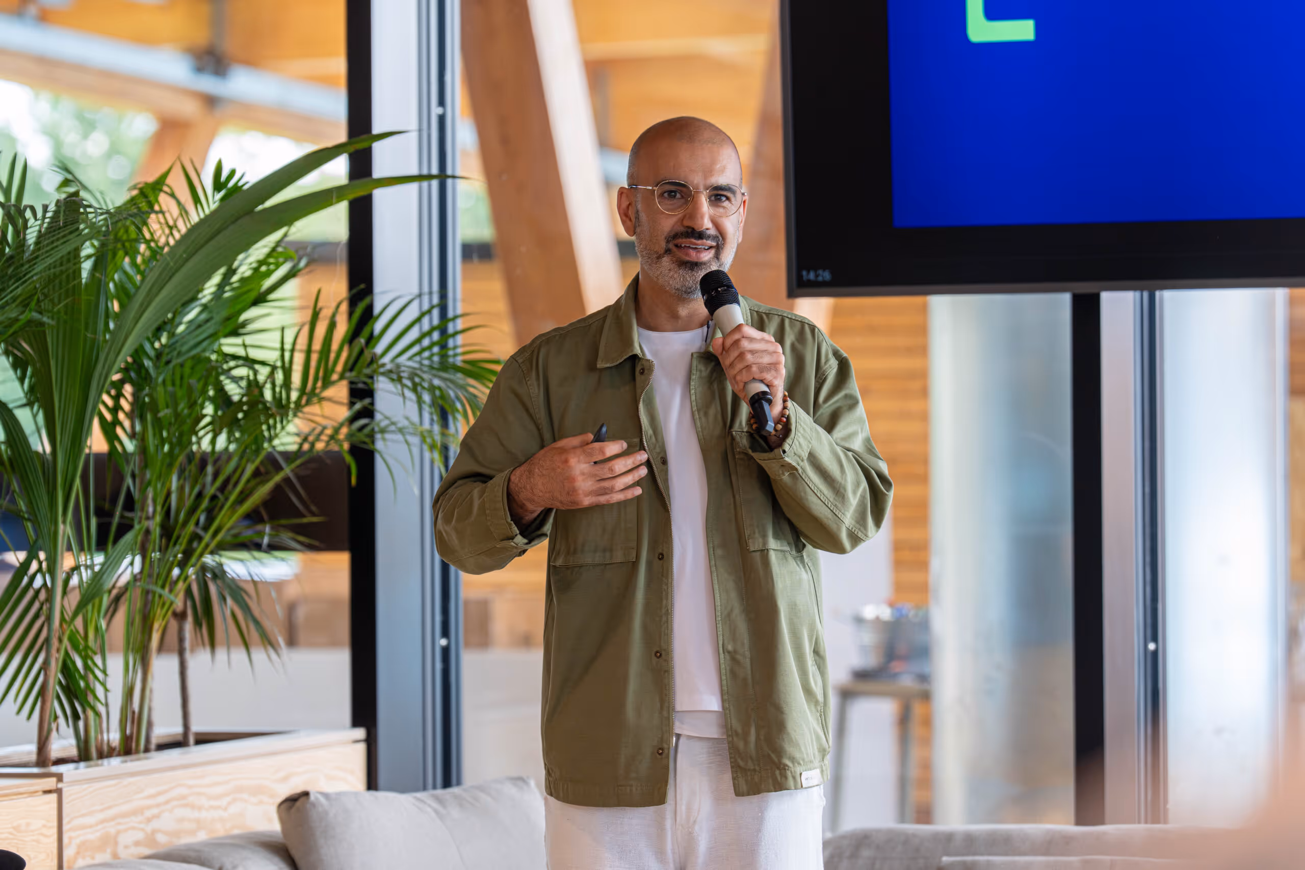 Man with glasses speaking into a microphone during a presentation in a modern room with plants.
