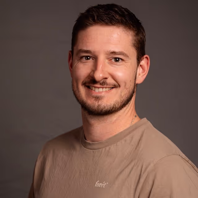 Smiling man with short brown hair and beard wearing a beige shirt against dark background.