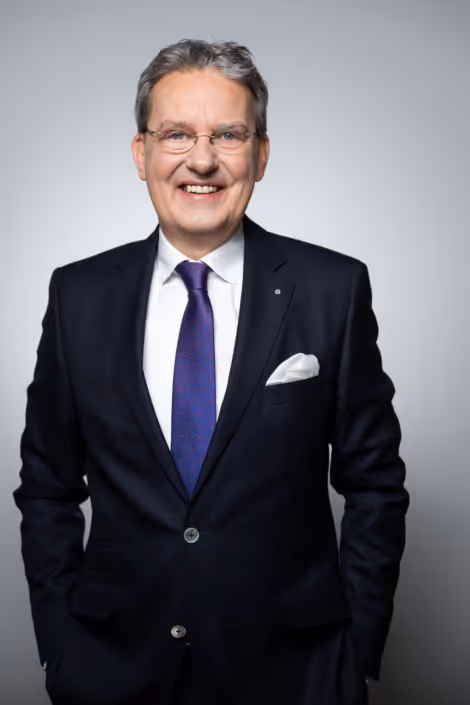 Smiling middle-aged man in glasses wearing a dark suit, white shirt, and purple tie with pocket square.