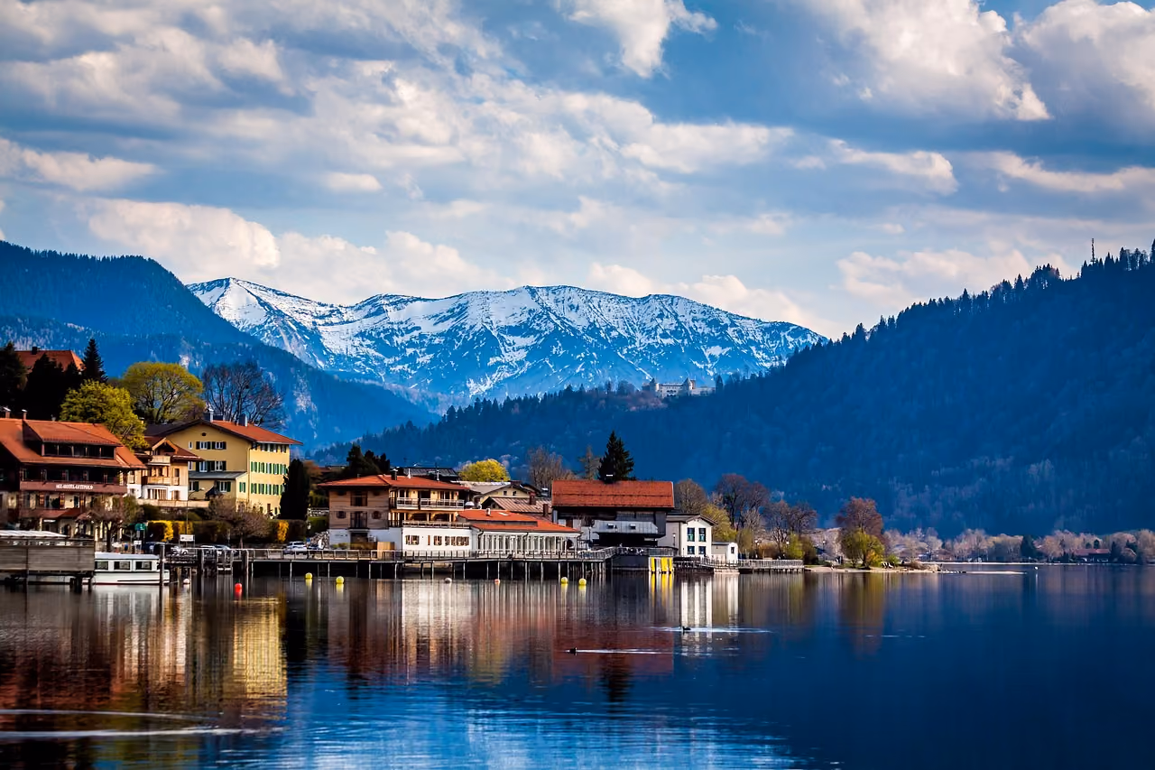 Lake with reflections, houses on the shore, forested hills, and snow-capped mountains under cloudy sky.