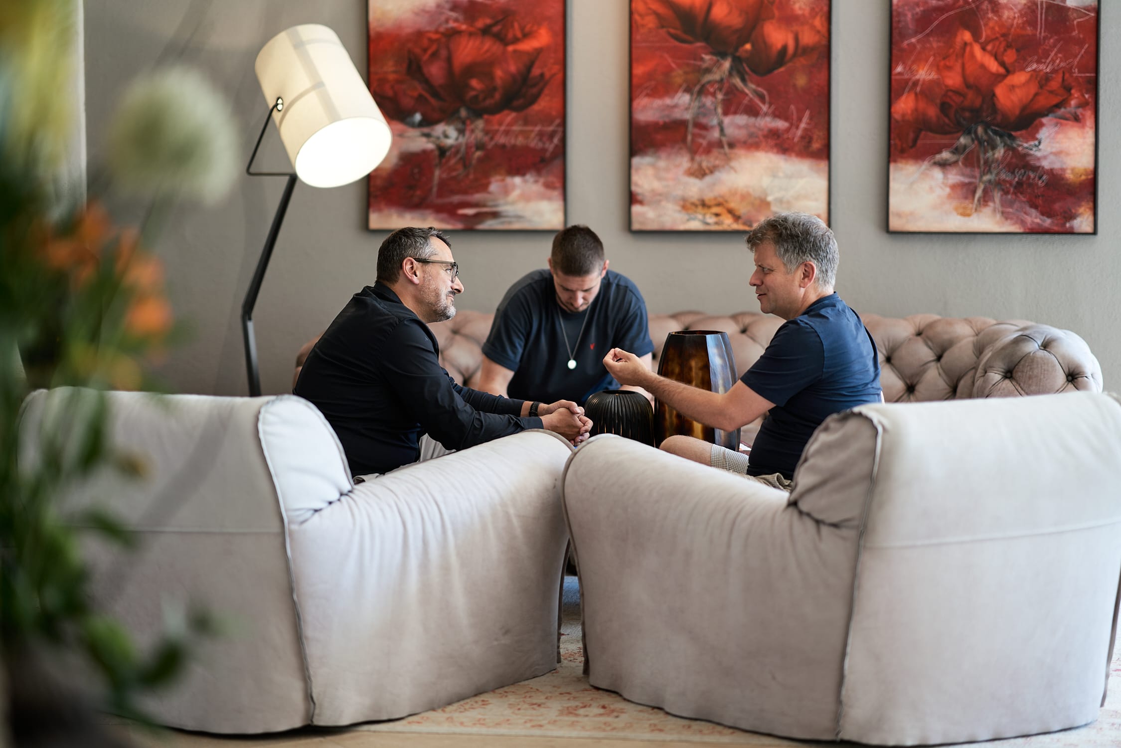 Drei Männer sitzen auf Sofas und einem Sessel, führen eine Unterhaltung in einem Wohnzimmer mit roten Gemälden.