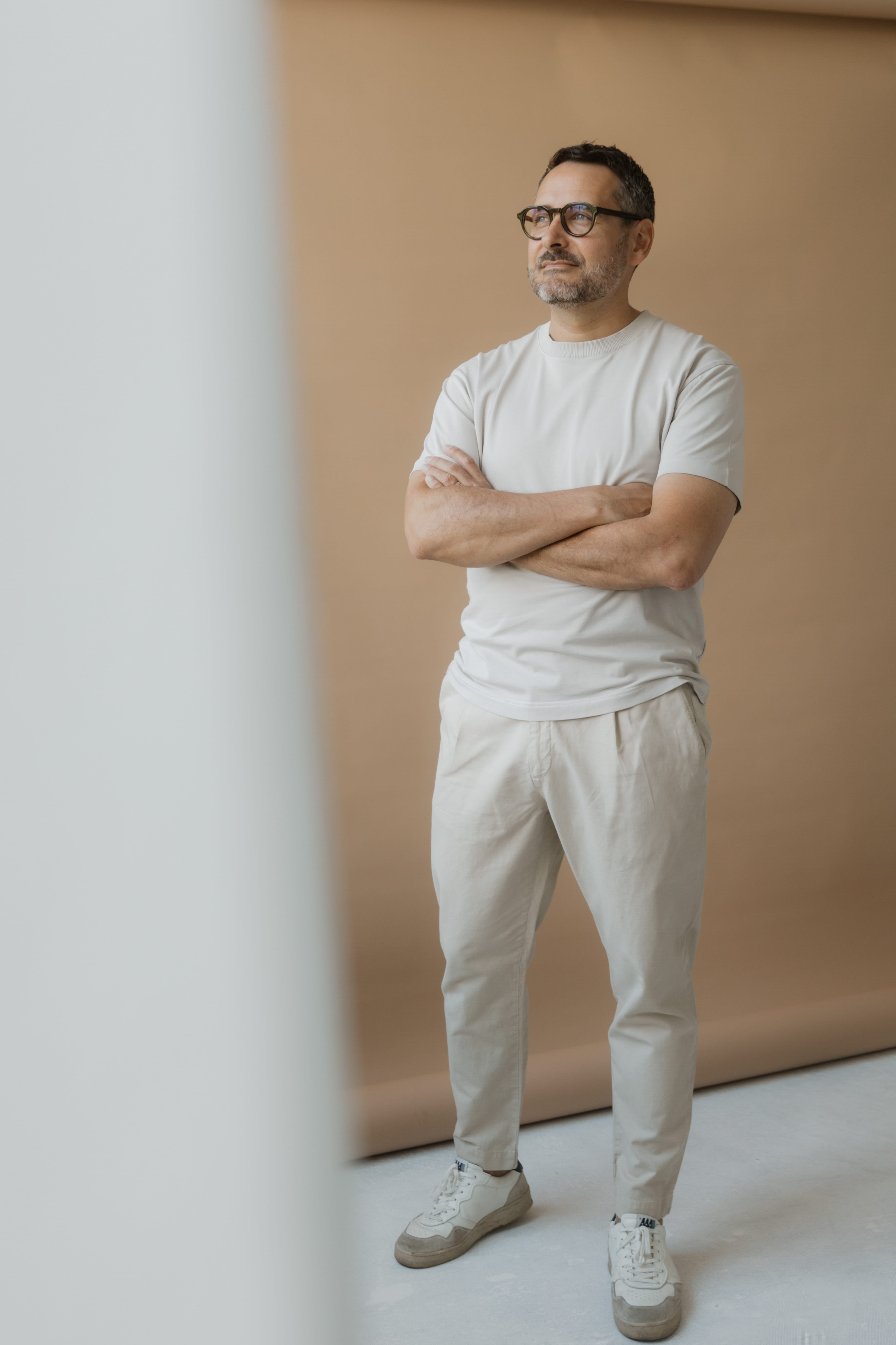 Mittelalter Mann mit Brille, kurzärmligem weißen Shirt und beigen Hosen vor braunem Hintergrund.