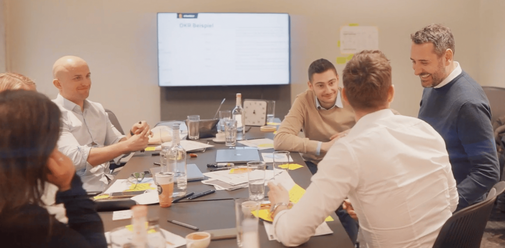 Fünf Personen unterhalten sich bei einem Meeting in einem Besprechungsraum mit Laptop und Notizen.