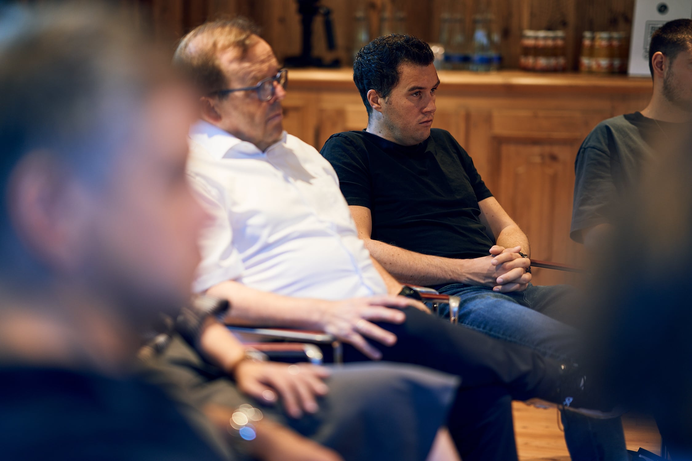 Mehrere Menschen sitzen aufmerksam in einem Raum mit Holzmöbeln und konzentrieren sich auf eine Diskussion.