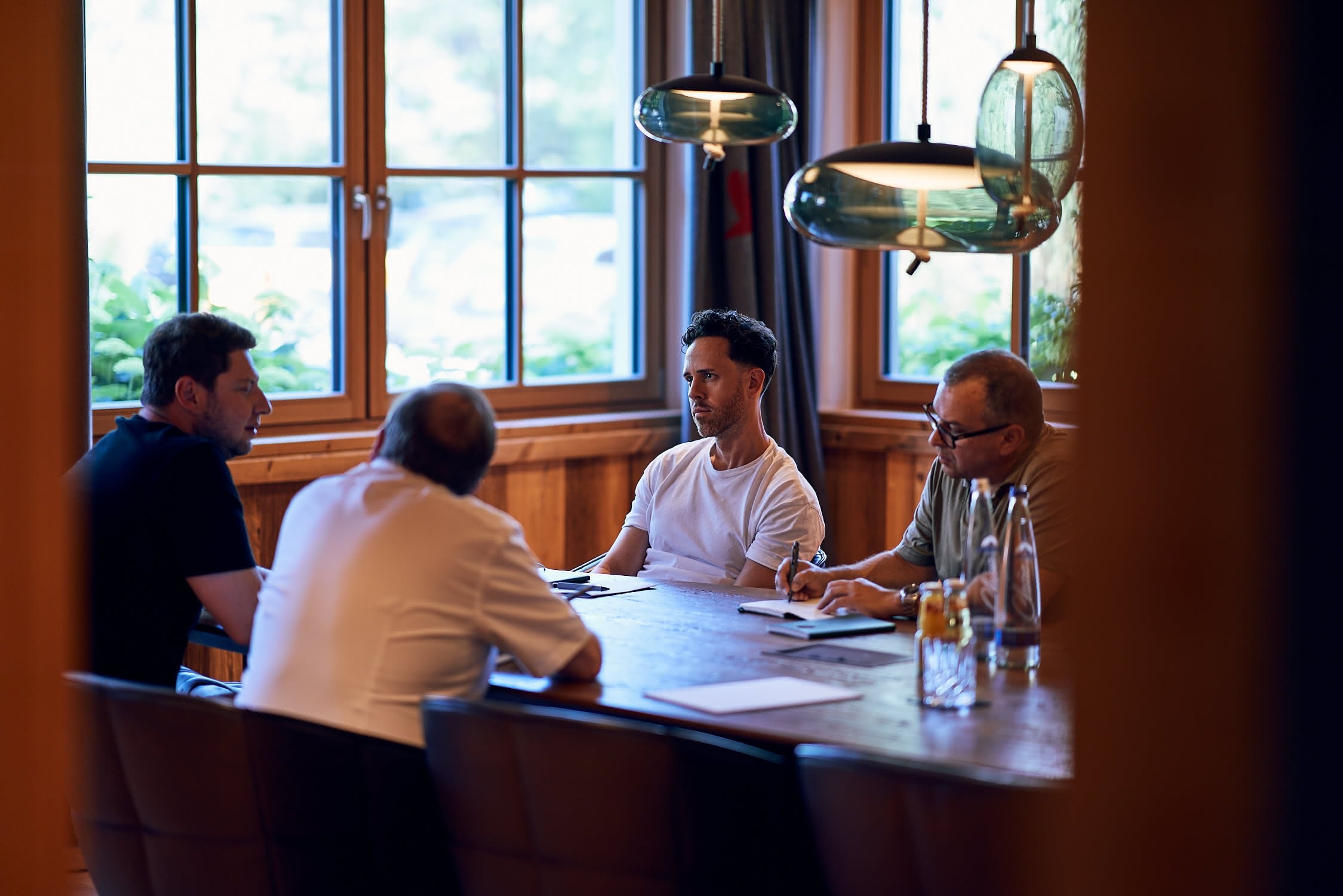 Vier Männer sitzen in einem Raum mit Holzvertäfelung und Fenstern, besprechen sich bei einem Tisch.