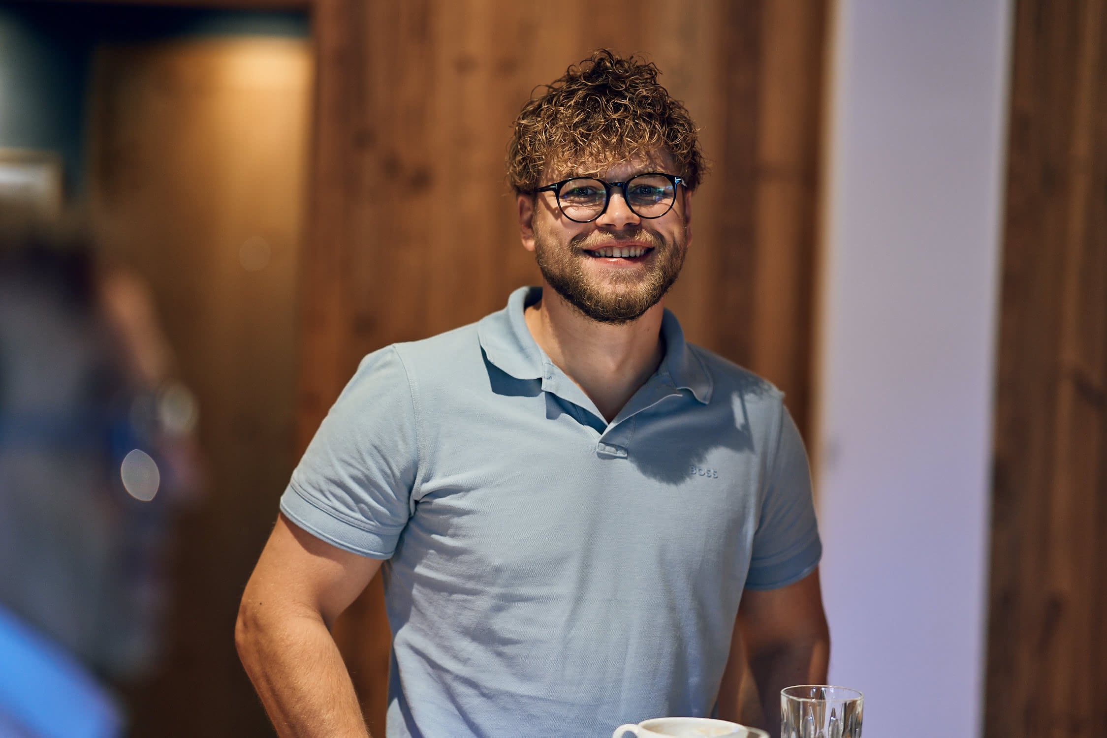 Junger Mann mit lockigem Haar und Brille lächelt in einem gemütlichen Raum mit Holzverkleidung.