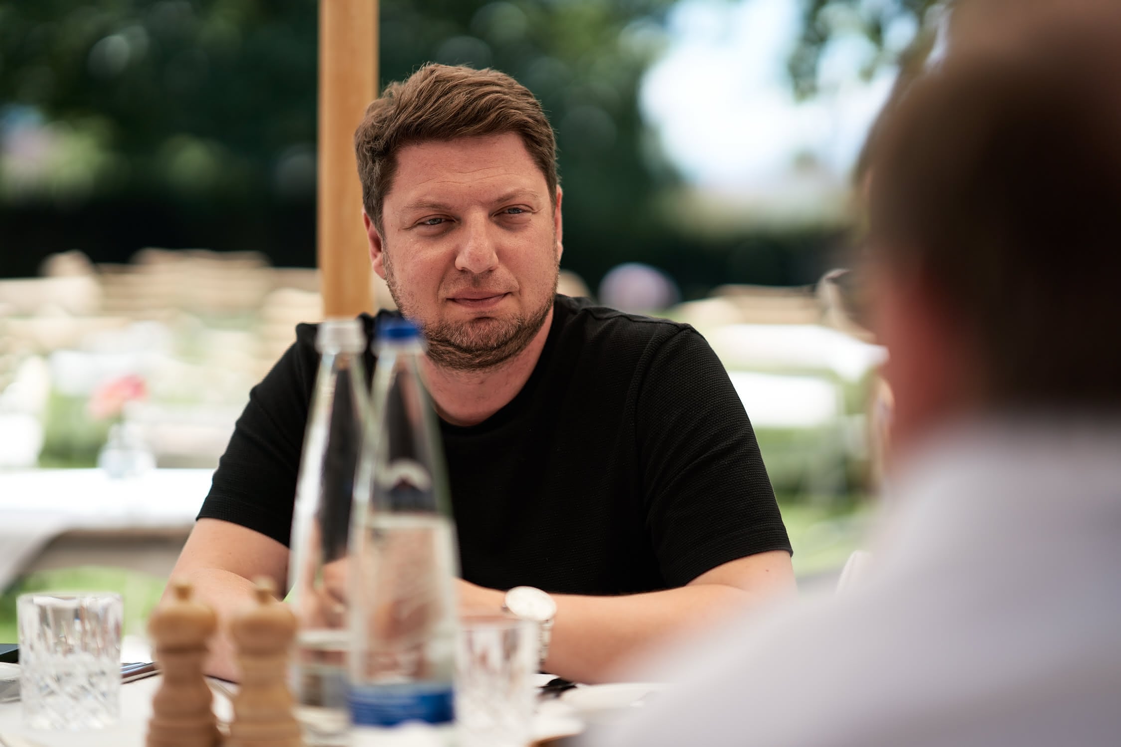 Mann in schwarzem T-Shirt sitzt an einem Tisch im Freien und blickt nach rechts.