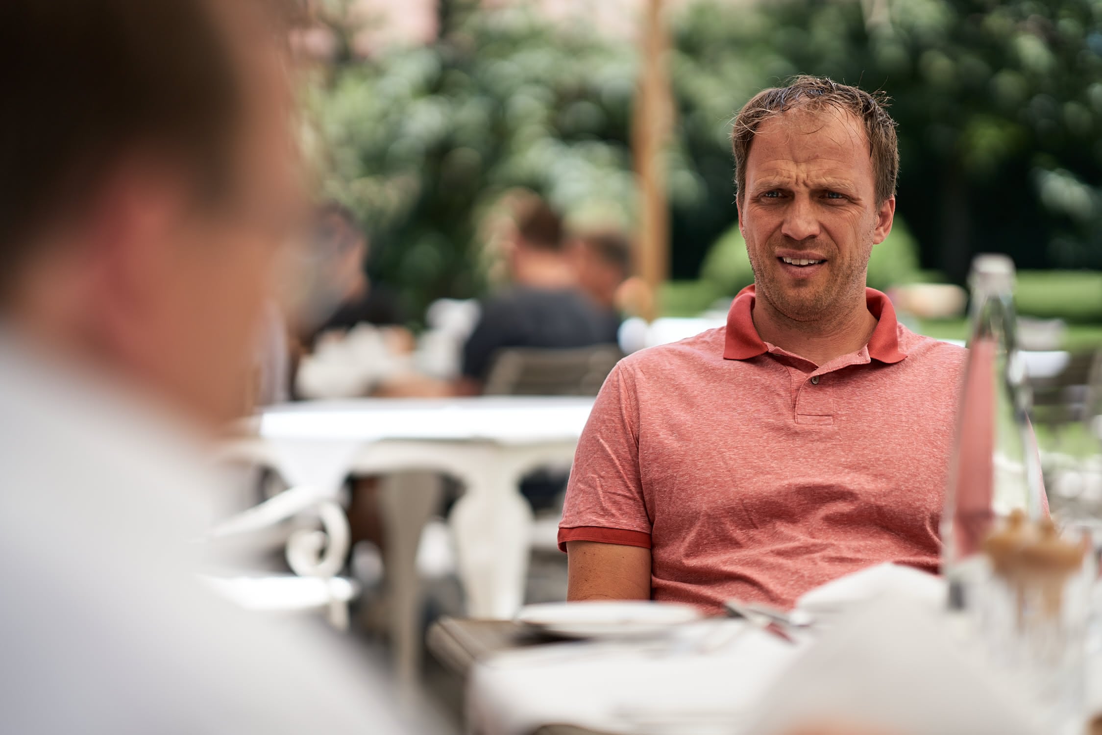Mann in rotem Poloshirt sitzt draußen bei Tisch und blickt skeptisch auf Gesprächspartner.