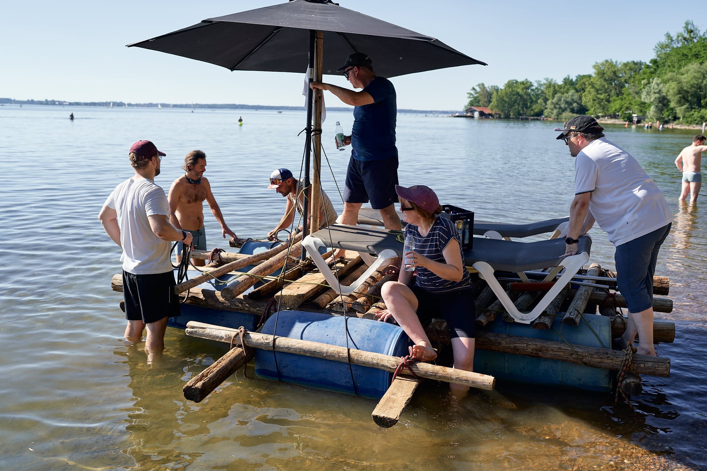 Sechs Menschen auf und um ein selbstgebautes Floß aus Fässern und Holz in flachem Seewasser.
