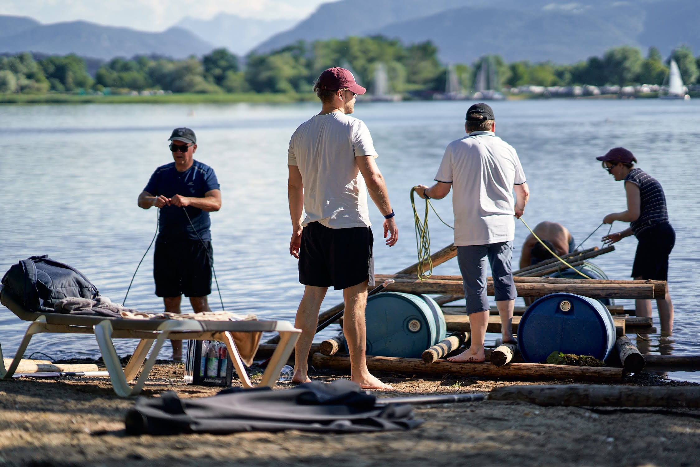 Vier Menschen bauen ein Floß aus Fässern und Holz am Ufer eines Sees mit Bergen im Hintergrund.