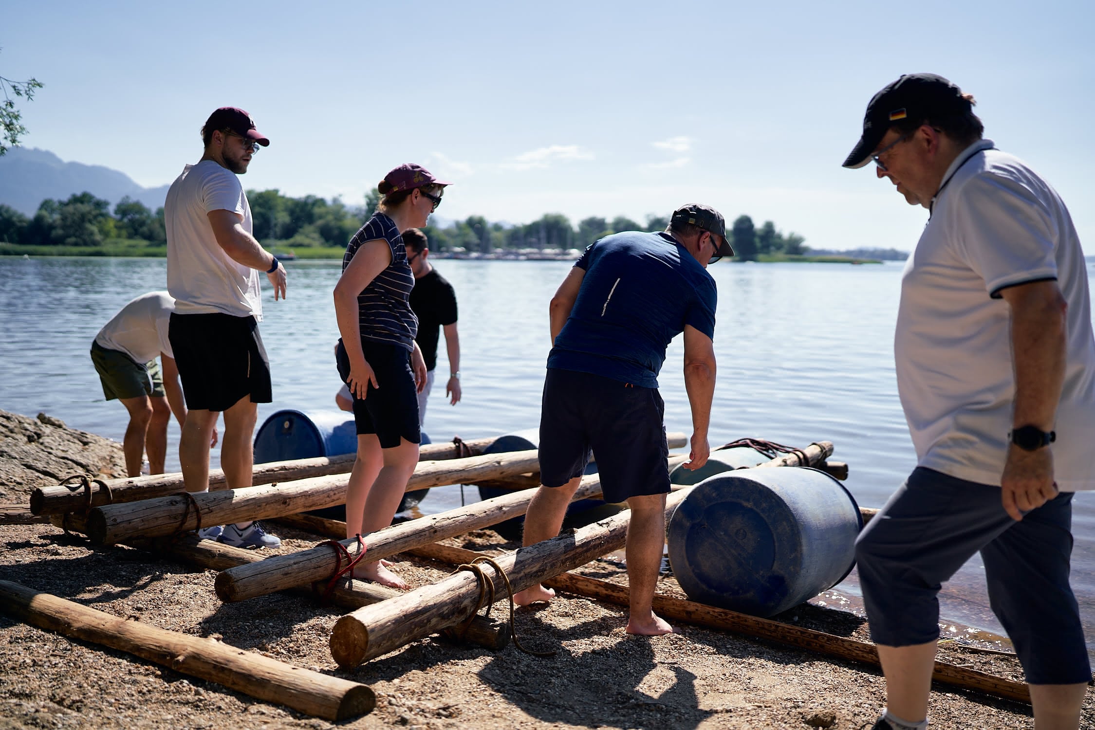 Sechs Menschen bauen ein Floß aus Holz und blauen Fässern am Ufer eines Sees.