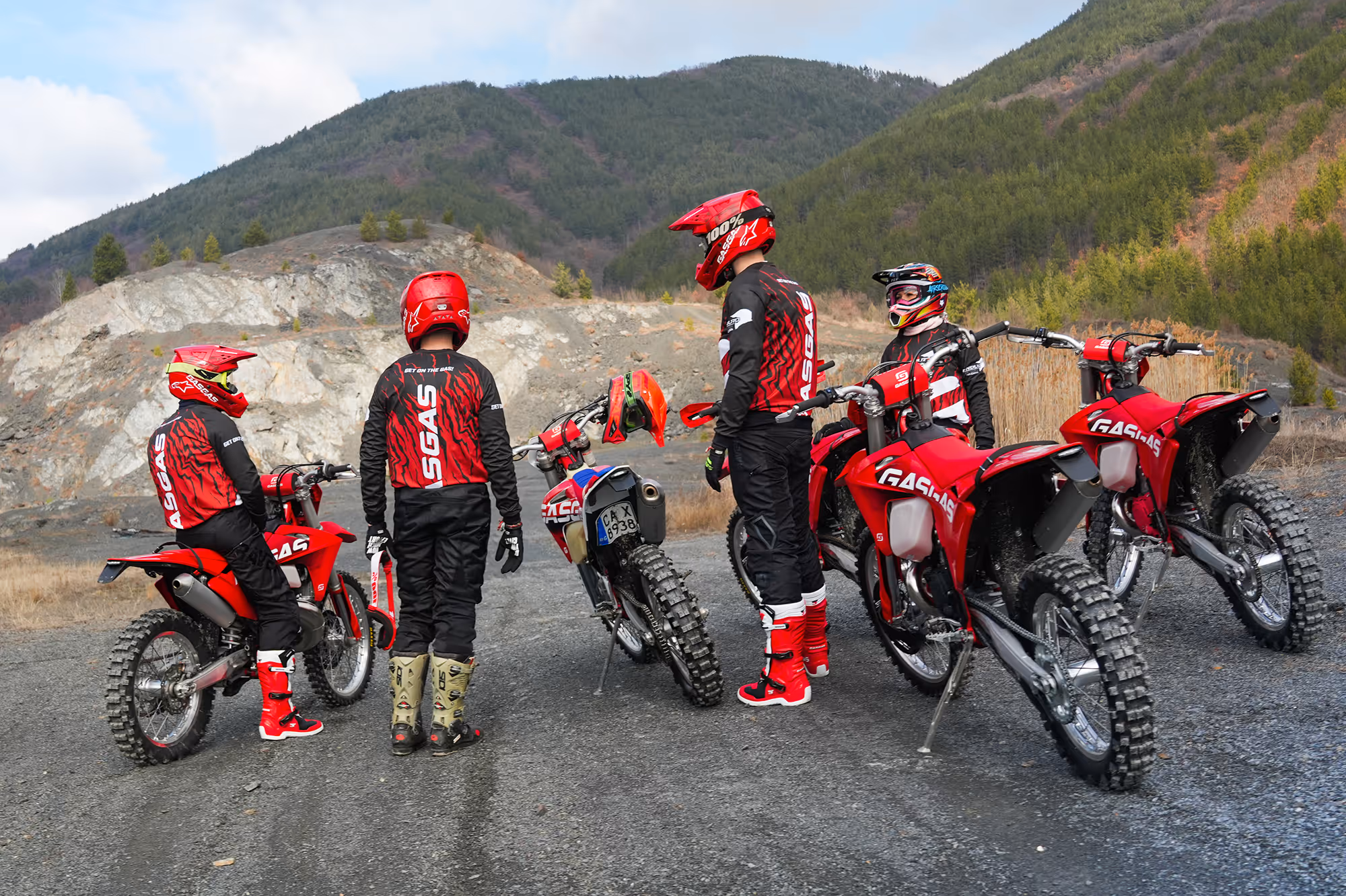 Four enduro riders wearing red helmets and gear stand next to four red enduro bikes on a gravel path with forested Bulgarian hills in the background.