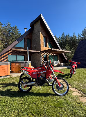 Two red enduro bikes parked on green grass in front of an A-frame cabin surrounded by trees.