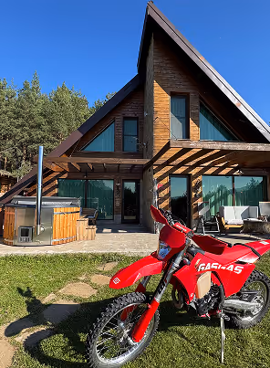 Red enduro bike parked on green grass in front of a wooden A-frame cabin with large windows and patio furniture under a clear blue sky.