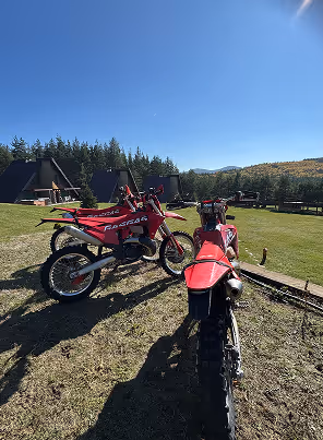 Two red enduro bikes parked on grass with A-frame cabins and trees under a clear blue sky.