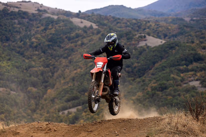 Motorcyclist in protective gear and helmet jumping a red enduro bike on a dirt trail with Bulgarian forested hills in the background.