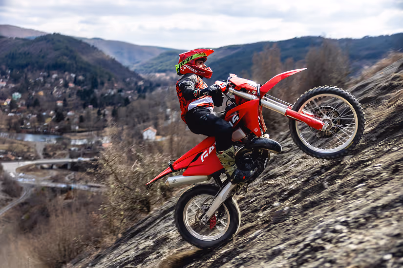 Person wearing a red helmet and gear riding a red enduro bike up a steep rocky hill with a blurred Bulgarian hillside town in the background.