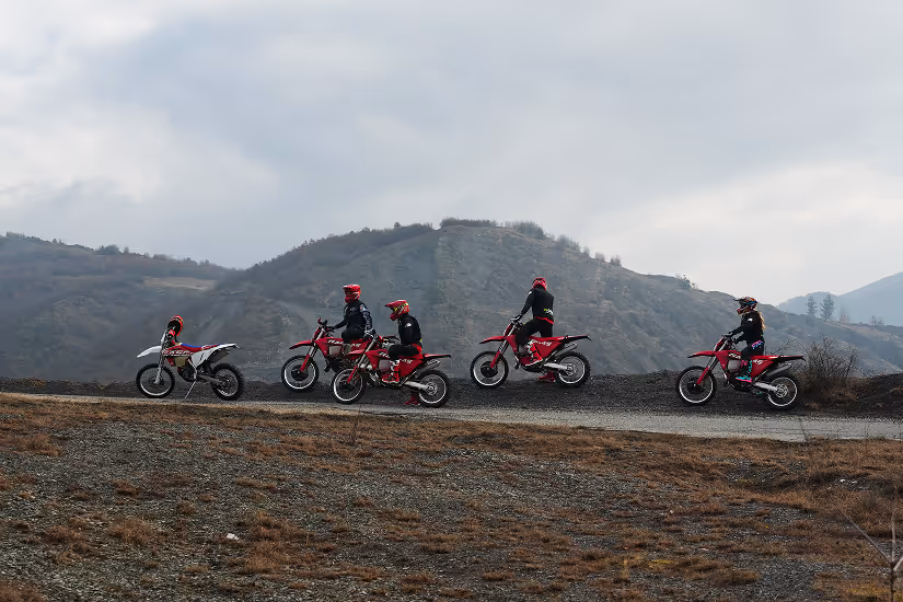 Five motorcyclists in red helmets riding enduro bikes on a Bulgarian gravel path with hills in the background.