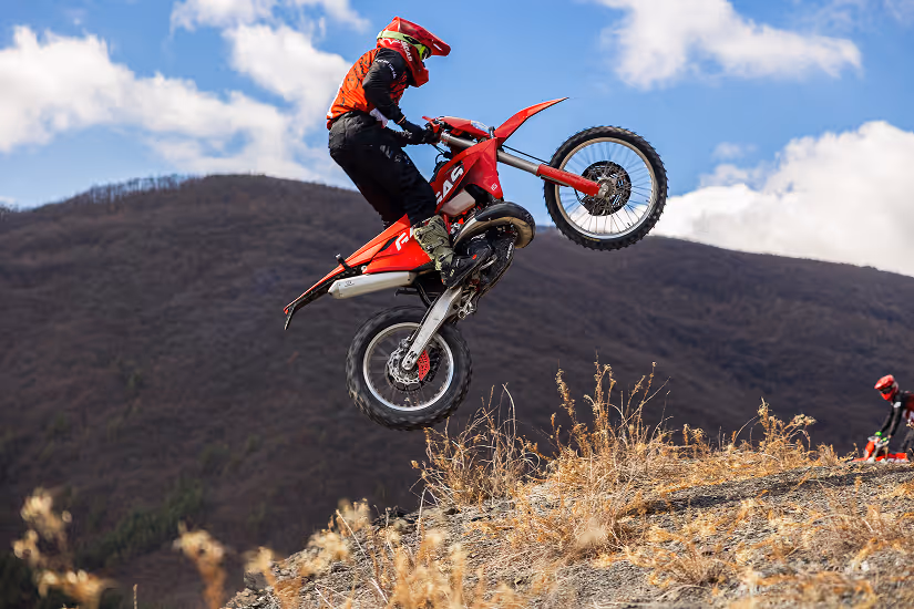 Person in red and black gear performing a jump on a red enduro bike over a rocky hill with Bulgarian mountains and blue sky in the background.