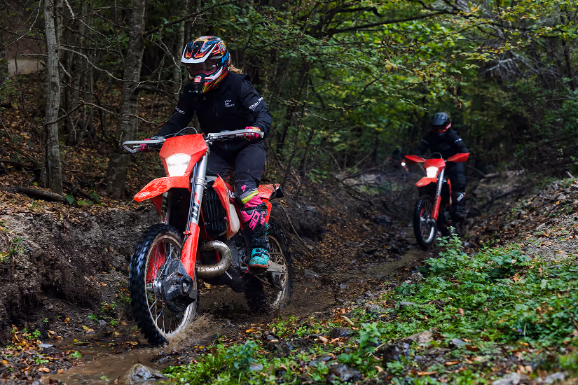 Two people riding red enduro bikes on a muddy Bulgarian forest trail surrounded by trees and greenery.