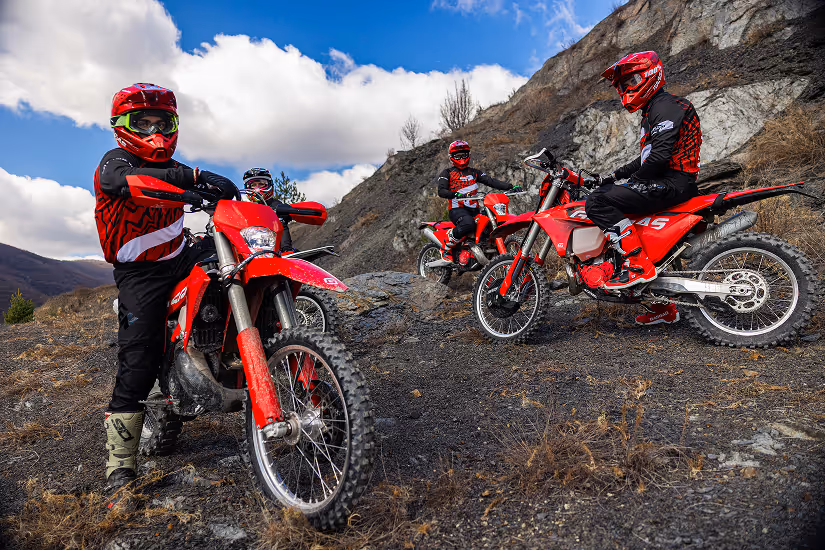 Three people in red helmets and gear riding red enduro bikes on a Bulgarian rocky hillside under a partly cloudy sky.