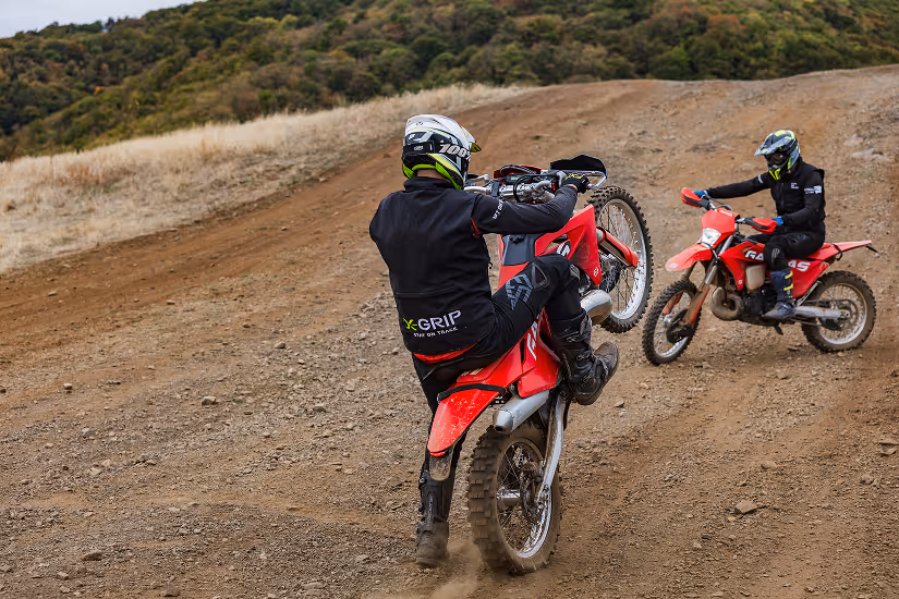Two motocross riders in black gear performing wheelies on red enduro bikes on a dirt trail with hills and Bulgarian greenery in the background.