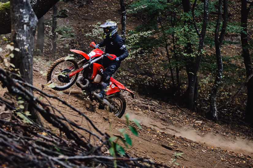 Person wearing black protective gear riding a red enduro bike uphill on a forest trail in Bulgaria.