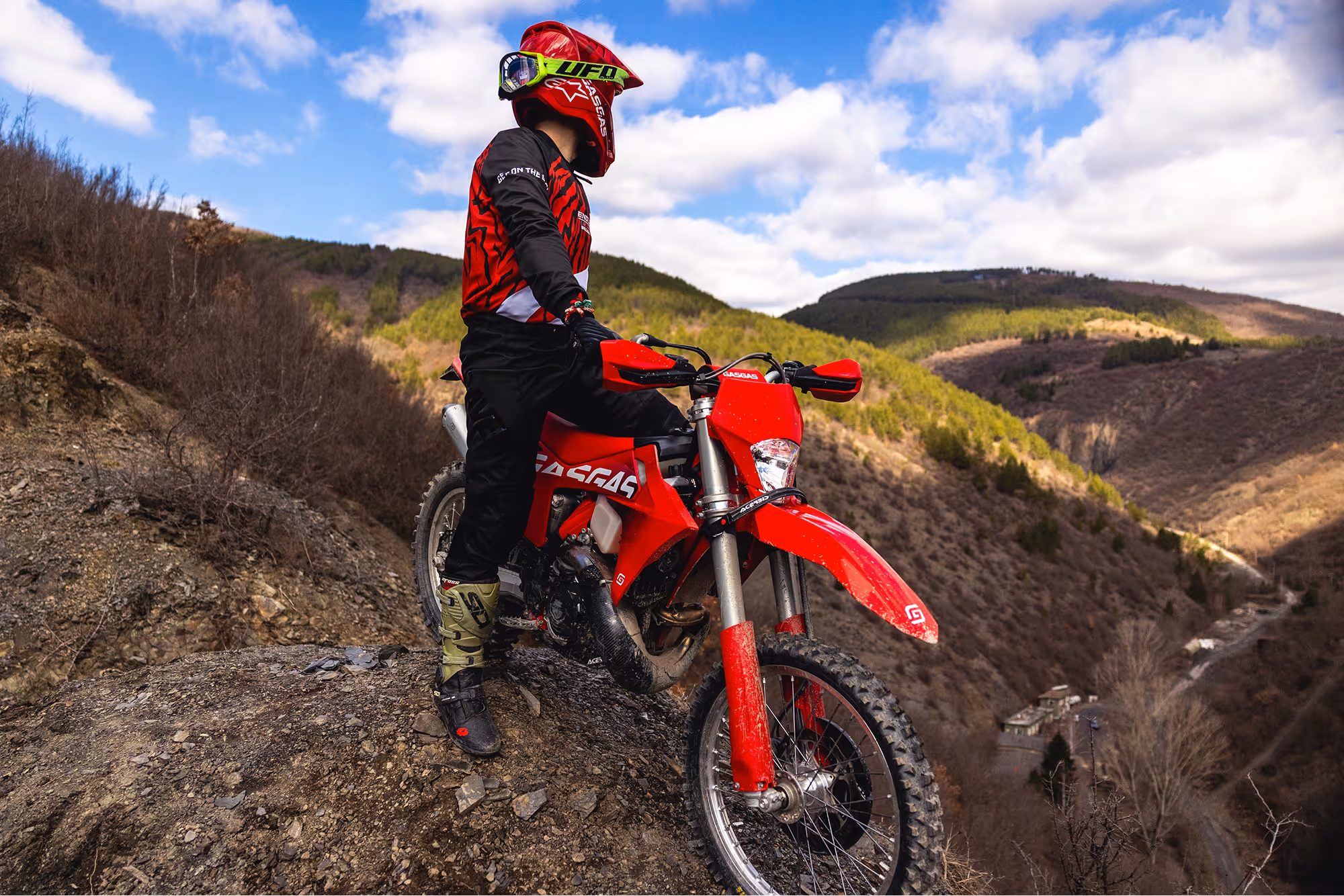 Motorcyclist in red gear standing with a red enduro bike on a rocky Bulgarian hill overlooking a mountainous landscape.