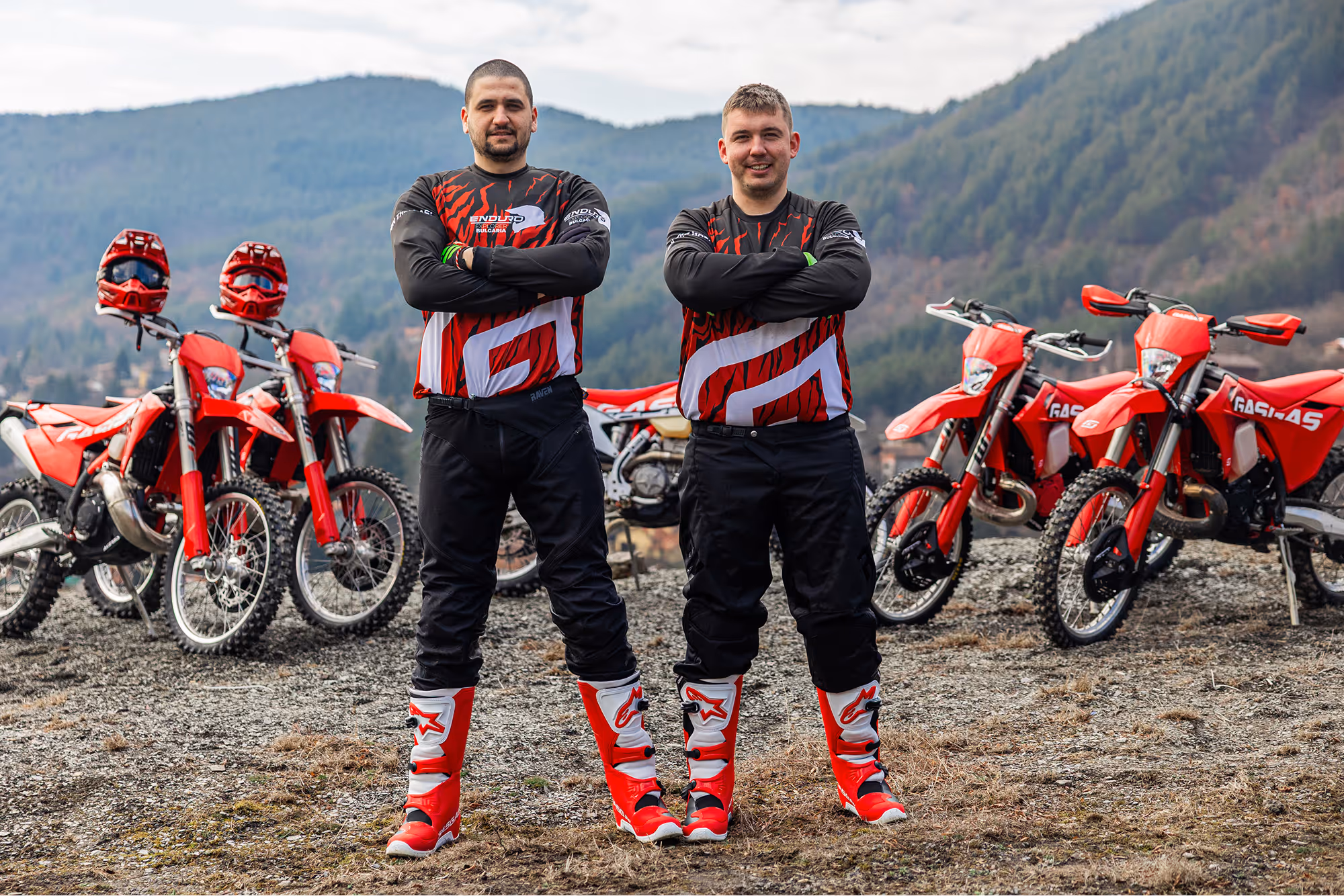 The two founders in matching red, black, and white motocross gear standing with arms crossed in front of five red enduro bikes and helmets, with Bulgarian forested hills in the background.
