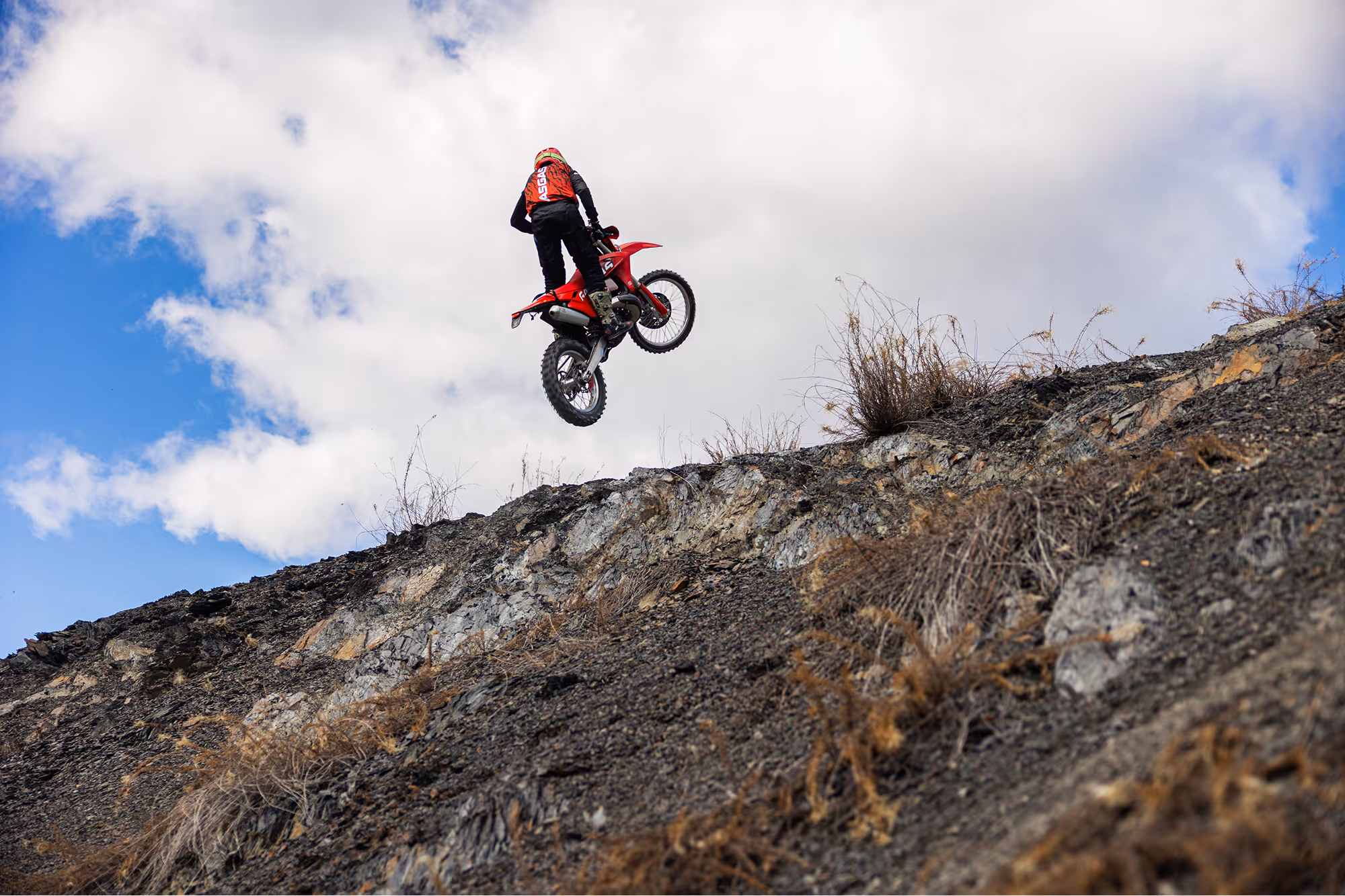 Motorcyclist in red gear performing a jump on a enduro bike over rocky Bulgarian terrain under a cloudy sky.