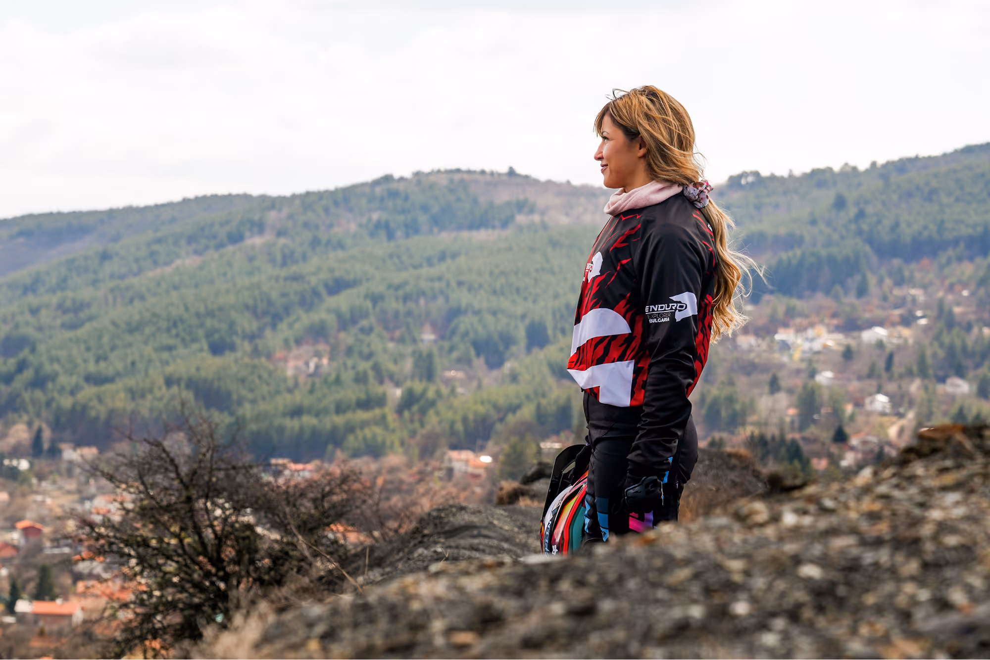 Woman in motorbike gear holding a helmet, standing outdoors with Bulgarian forested hills in the background.